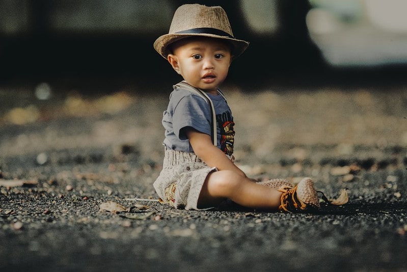 Baby boy wearing dungaree shorts and a straw hat sitting on the floor on the street.