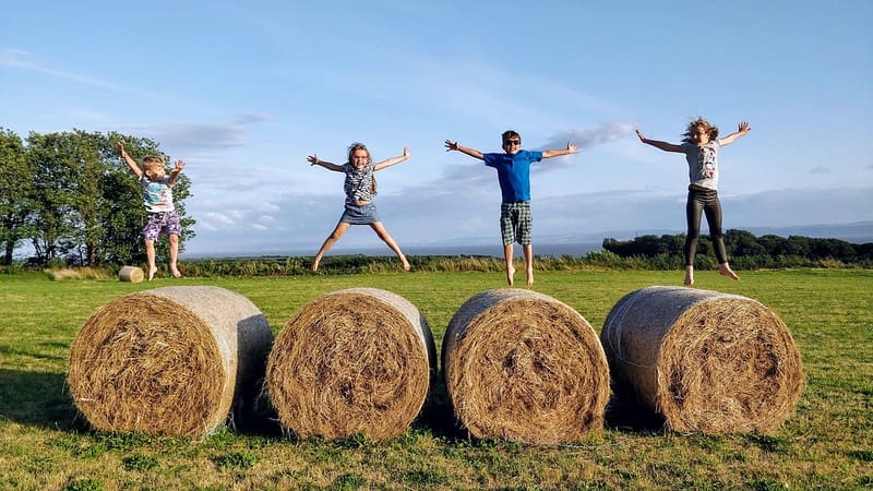 Four children jump on top of bales of hay, with the sea in the background