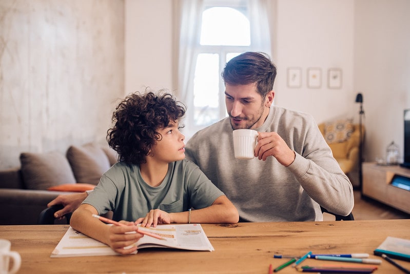 Schoolboy sat at table doing homework with help from his dad.