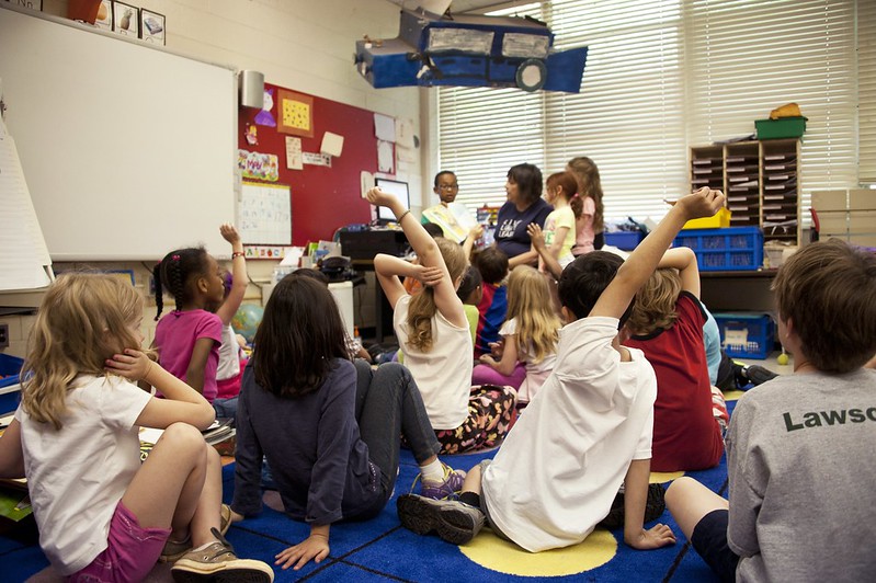 Classroom of kids raising their hand and sitting on the floor.