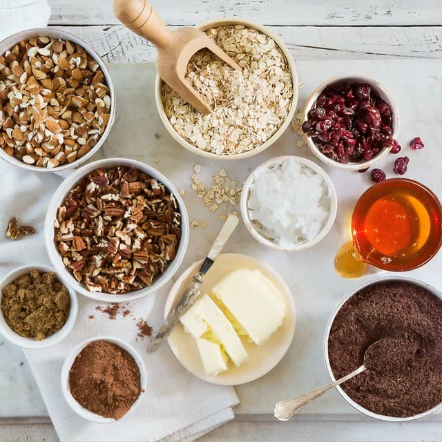 Various ingredients for flapjacks in bowls on a white wooden tabletop.