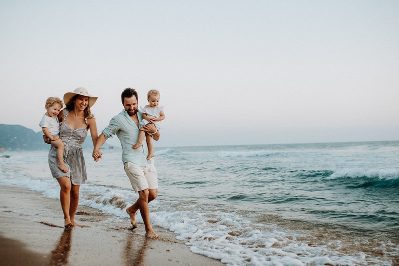 Mum and dad holding hands walking along the beach on holiday with babies in arms.