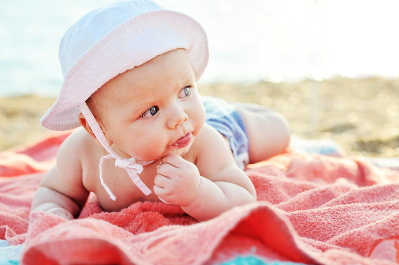 Baby wearing a sun hat lying on a pink towel on the beach on holiday.