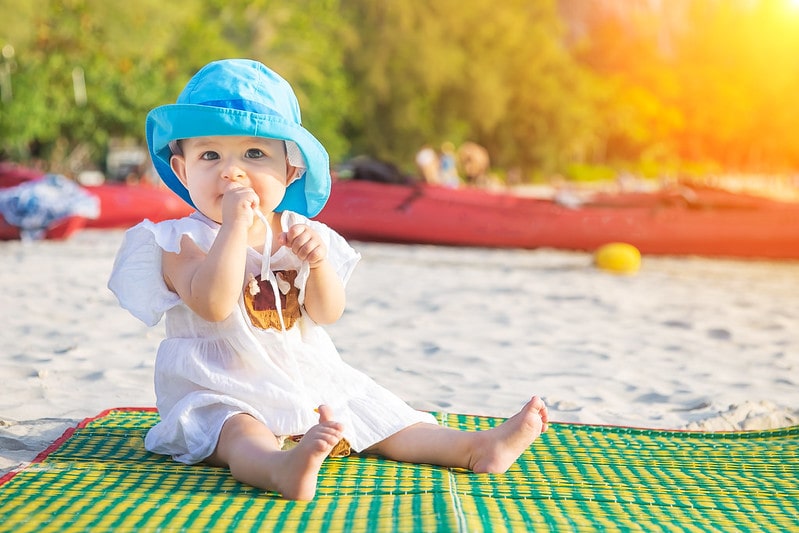 Baby wearing a blue sun hat sat on a rug on the beach on holiday.