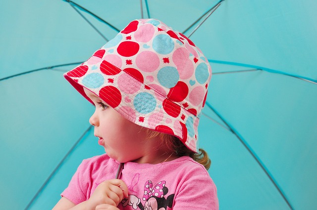 Girl wearing a colourful spotty sun hat holding a blue umbrella on holiday in the UK.