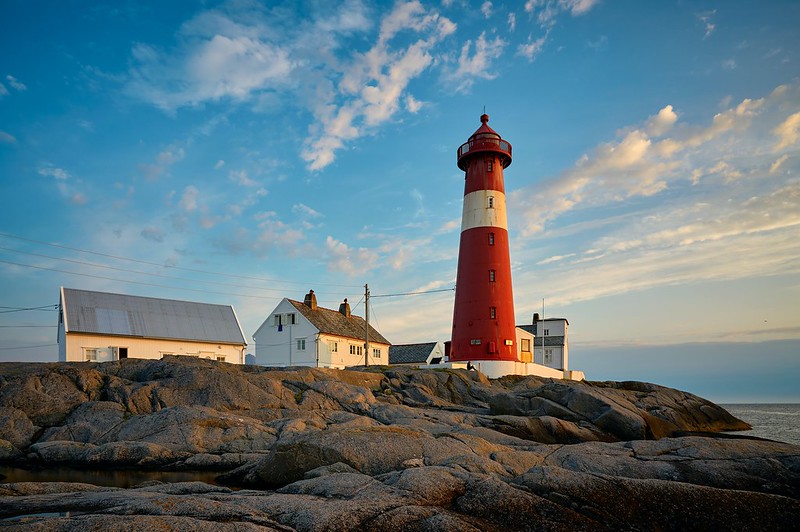 Red lighthouse against a slightly cloudy sky, with a white stripe. There are three houses in the background and a rocky shoreline in the foreground.