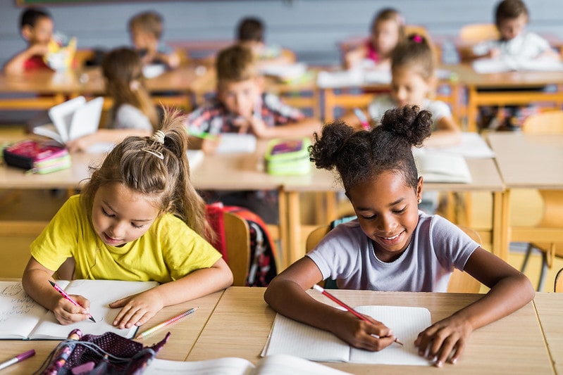 Two schoolgirls and their classmates sat at their desks learning multi-clause sentences.