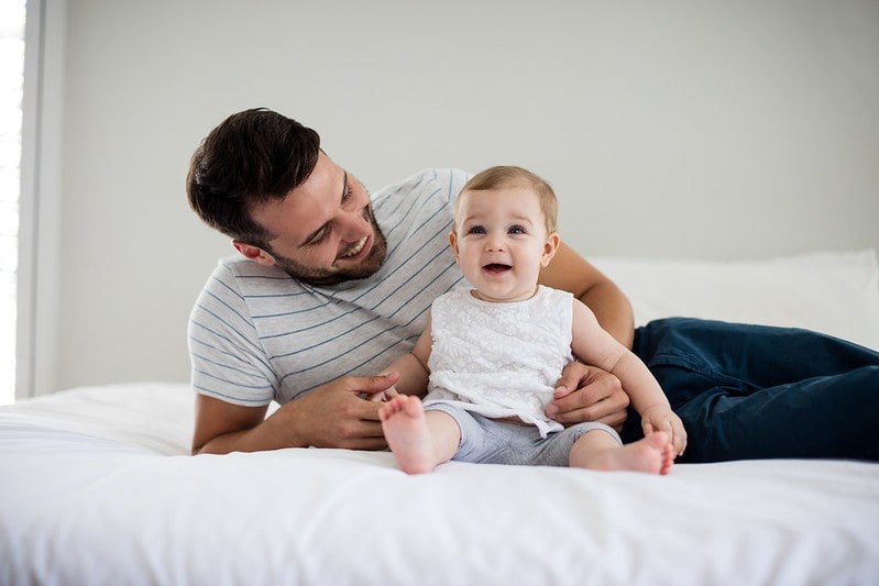 Dad sitting on bed smiling at his baby girl who is sat in his arms smiling.
