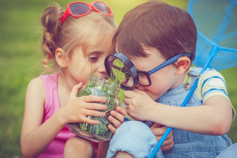 Two small children looking at a jar full of grass through a magnifying glass