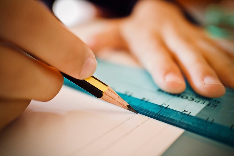 Close-up of someone trying to draw a straight line with a pencil and ruler. Their fingers, the pencil, the ruler, and the paper are in view.