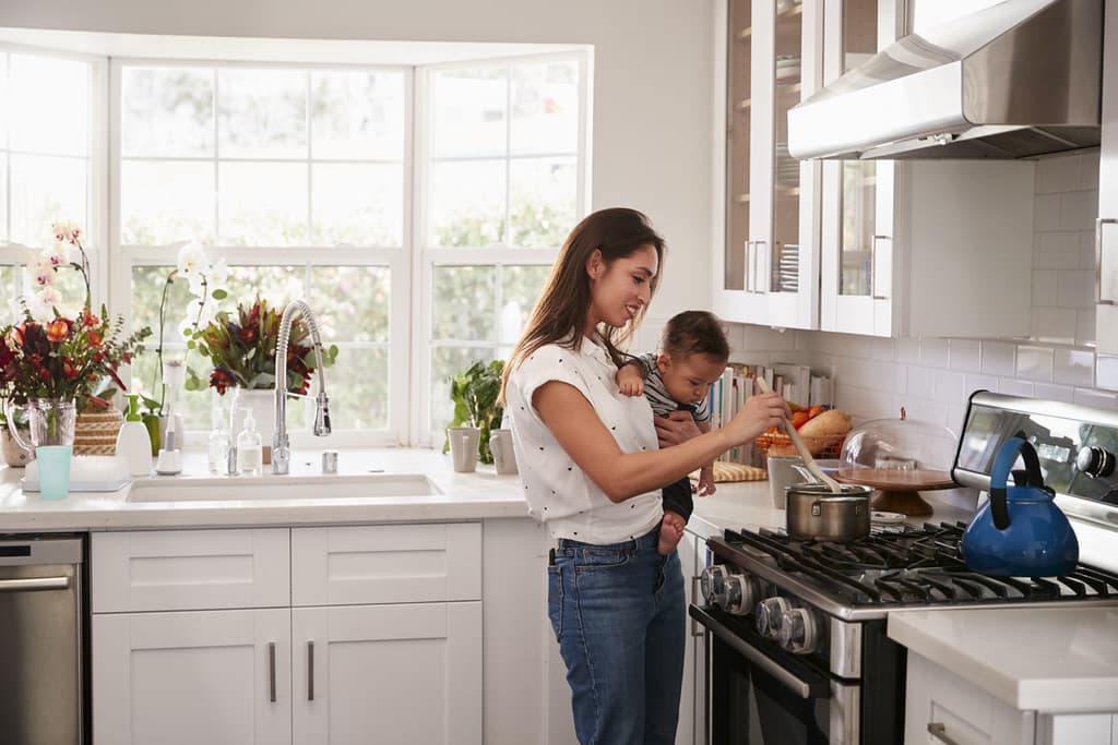Mum in kitchen stirring a pot while holding her baby in her arms.