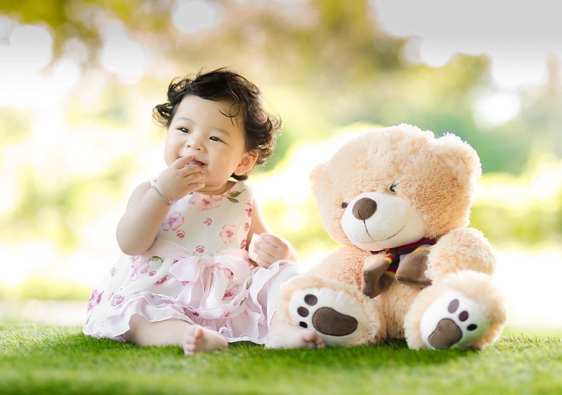 Baby girl in floral dress sat on the grass smiling next to her teddy bear.