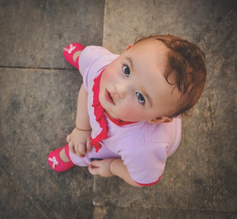 Baby girl wearing a pink outfit and pink shoes looking up at the camera.