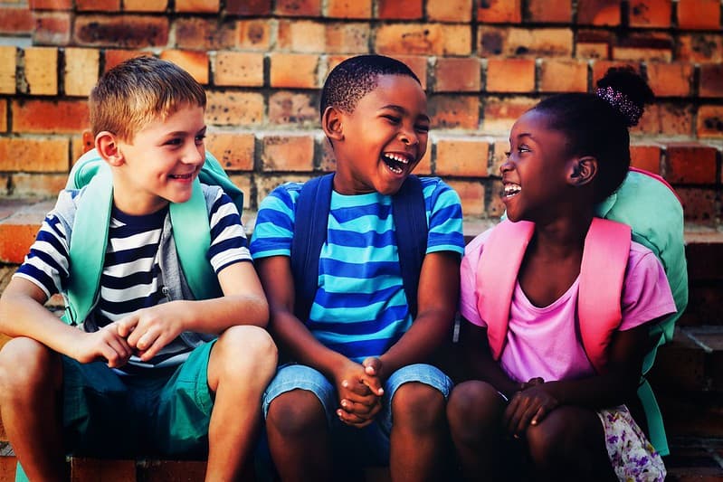 Three school kids sat by a wall laughing at jokes together.