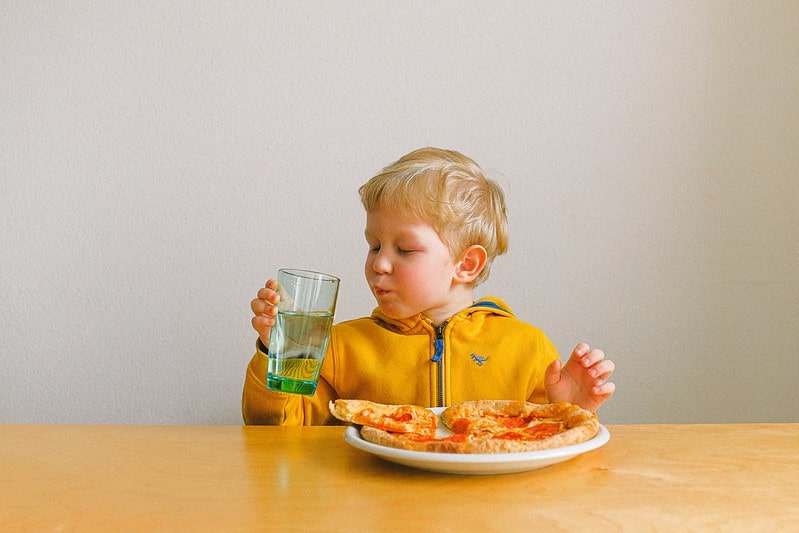 Young boy sat at the table eating pizza and drinking water.