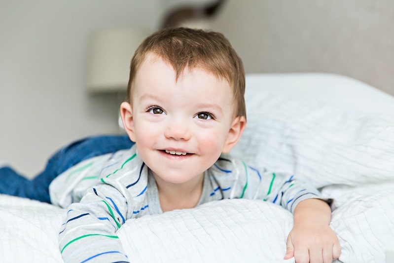 Little boy lying on his tummy on the bed looking up and smiling at his parents.l