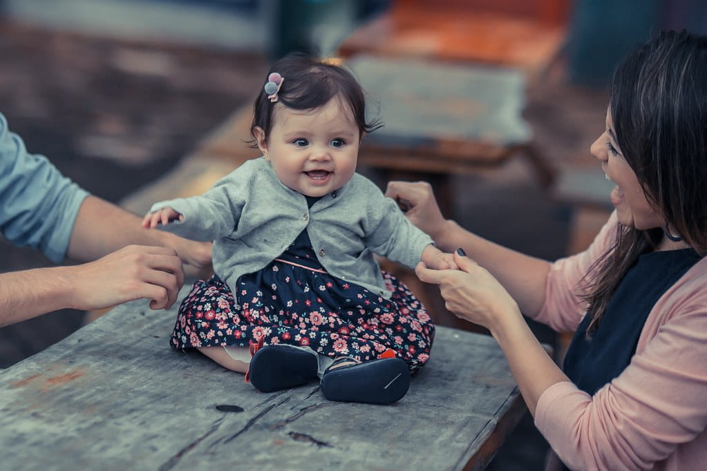 Baby girl in a floral dress and grey cardigan sat on a table smiling with her parents.