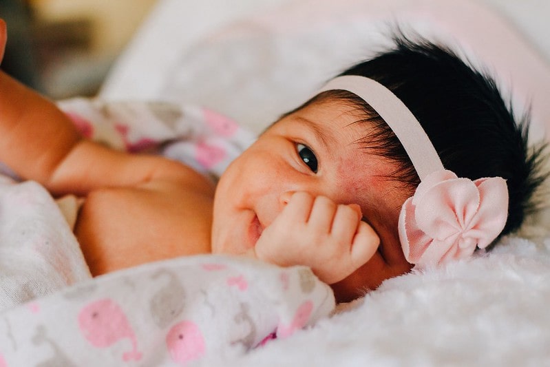 Baby girl wearing a flower hairband lying on the bed smiling, covering one eye with her hands.