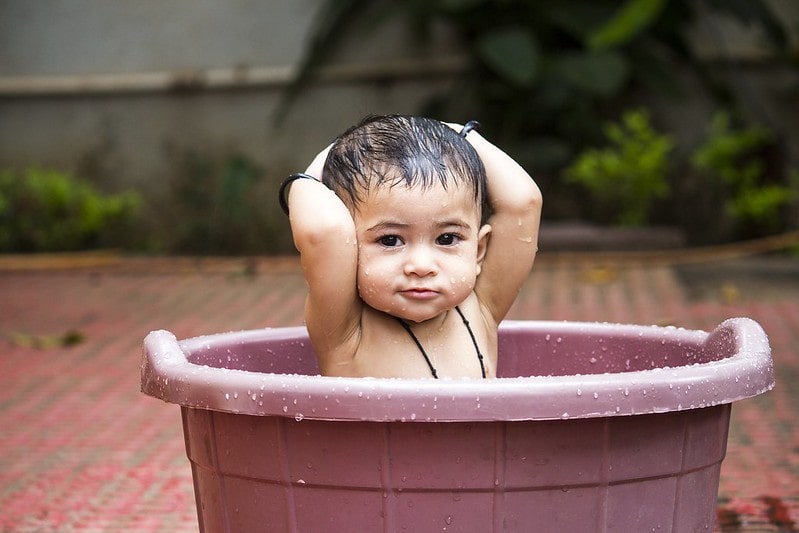 Baby girl sitting in a tub of water with her hands on her head.