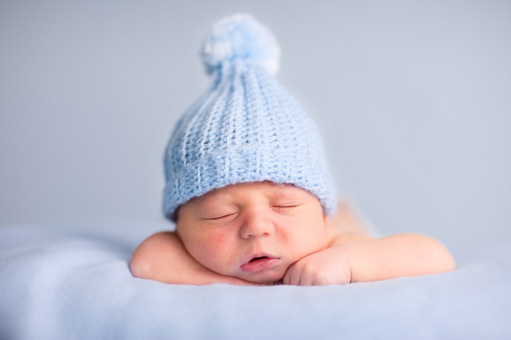Baby boy wearing a light blue woolly hat sleeping on his tummy.