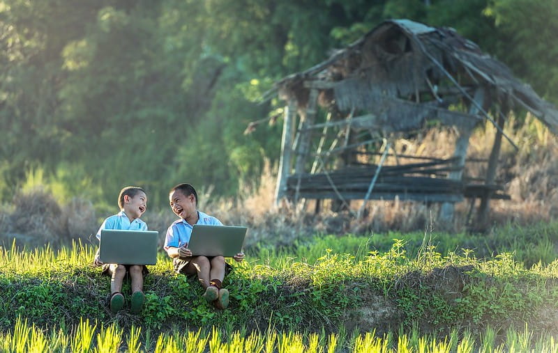 Two young boys sitting outside on the grass with laptops, both laughing.