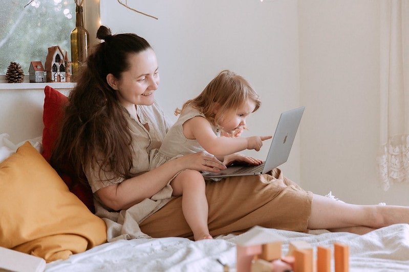 Little daughter sat on mum's lap on the bed looking and pointing at the laptop.
