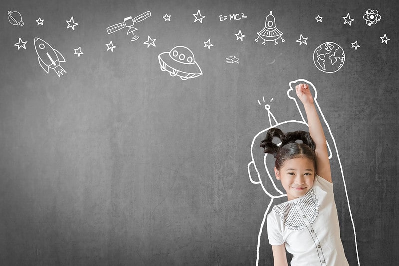 Little girl standing in front of an outline of an astronaut suit drawn on a blackboard with stars and spacecrafts drawn above.