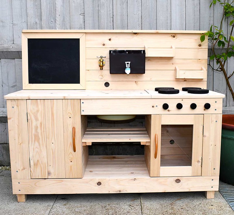 A Big Game Hunters Triple Mud Kitchen outside in the garden, with a working tap with hose attachment, large chalkboard, moveable draining board, opening oven, moveable dials, cupboard, and parcel shelf.