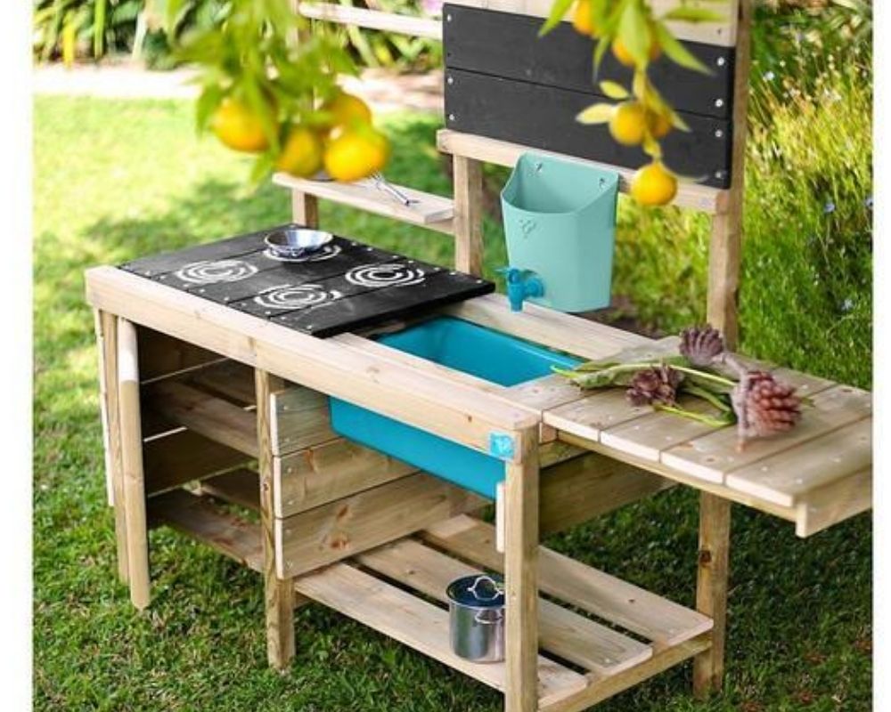 A TP Muddy Madness Wooden Mud Kitchen with removable tap, 4 hobs and DIY stencils, pans, whisk, blackboard, and extendable cooking area, placed outside in the garden.