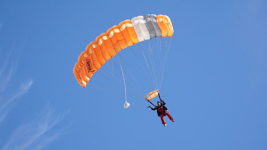 Man skydiving with an orange parachute is being pulled back down to the ground by gravity.