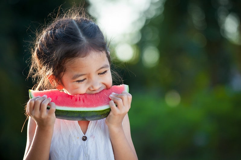 Girl biting into a wedge of watermelon, standing in the garden.