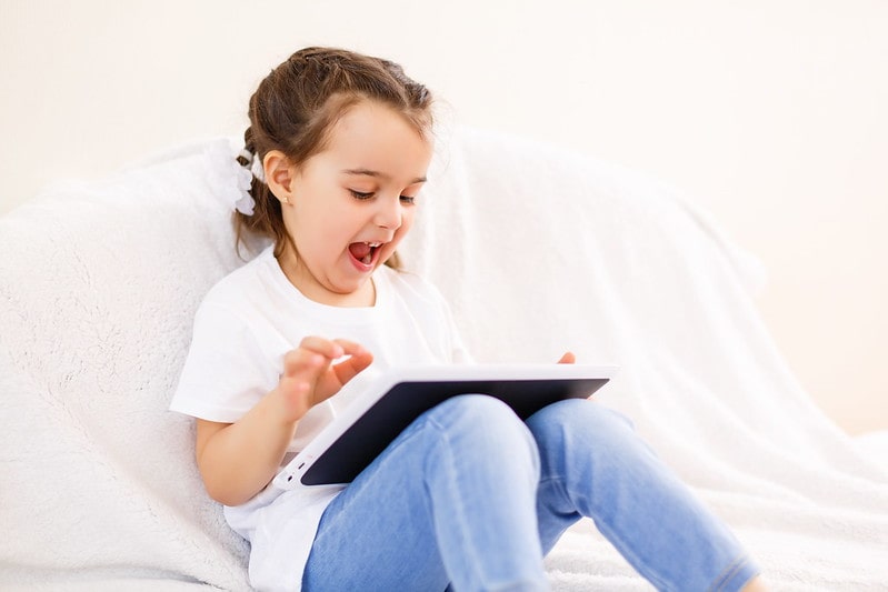 Little girl sat on the bed looking at computer jokes on her tablet and laughing.
