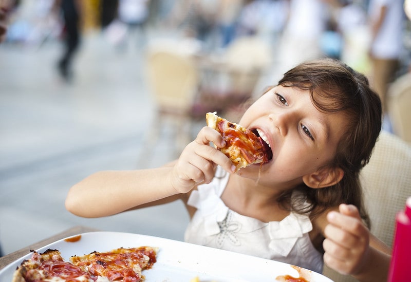 Little girl sat at the table in a restaurant eating a slice of pizza.