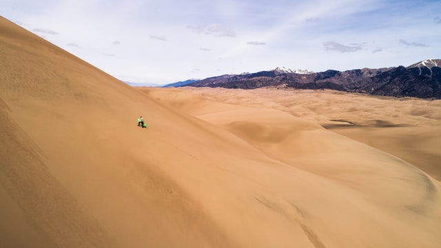 Man climbing up a large sand dune in Peru, mountains visible at the edge of the sand.