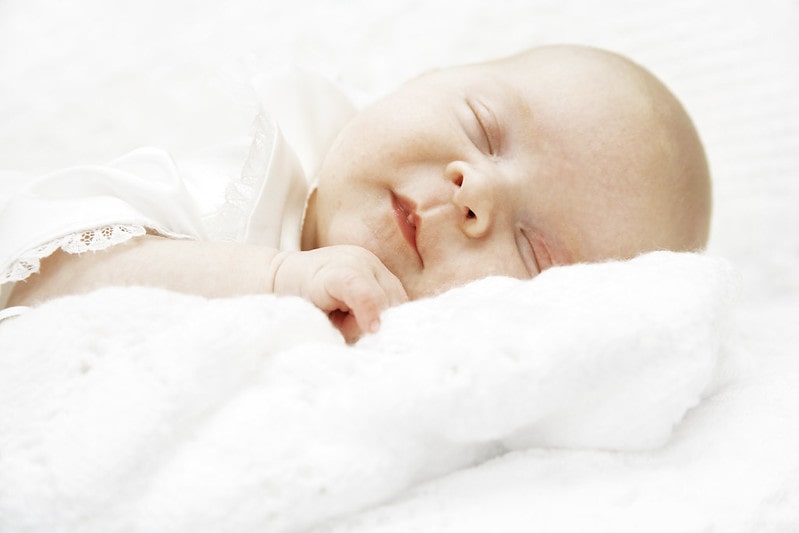Newborn baby dressed in white, sleeping on a white bed.