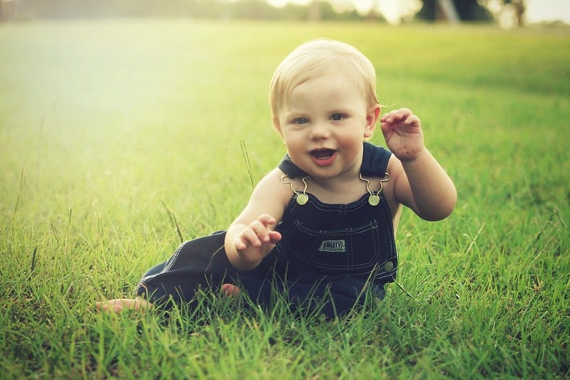 Blonde baby wearing dungarees sat in the grass smiling.