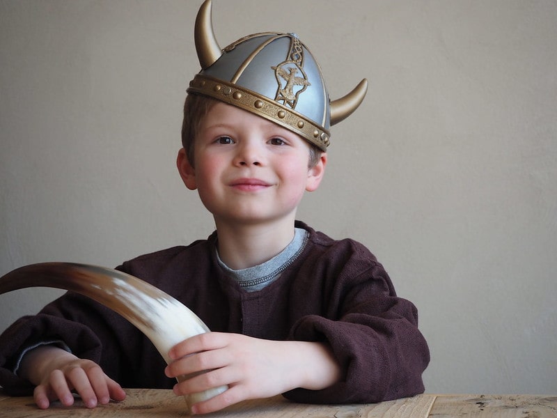 Young boy sat at a wooden table wearing a Viking helmet and holding a horn.