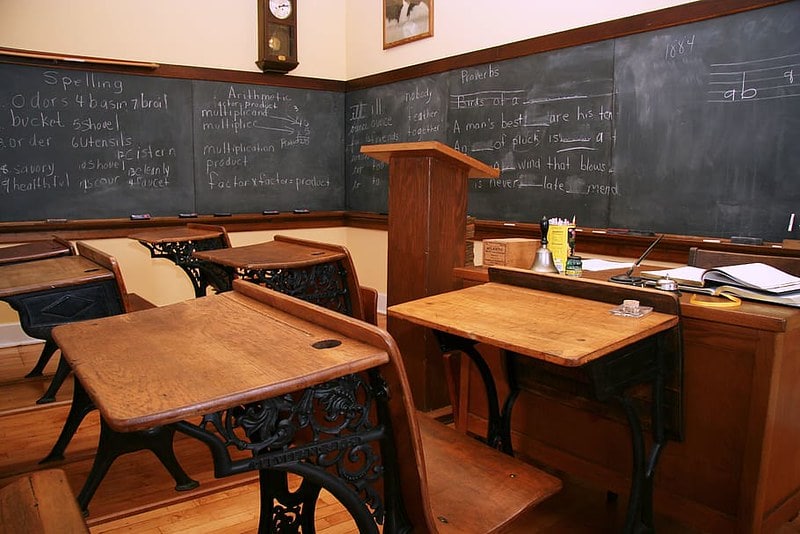 Victorian-era schoolroom with wooden desks and a blackboard.
