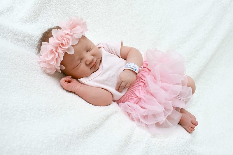 Baby girl wearing a pink tutu and matching flower crown, lying on a white blanket fast asleep.