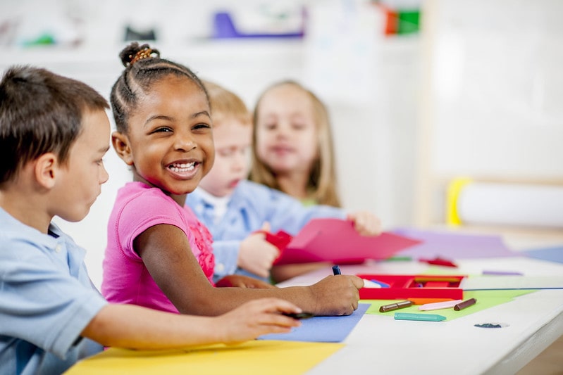 Kids sat at a table smiling, colouring on pieces of coloured paper.
