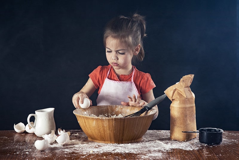 Little girl wearing a pink apron baking a cake, breaking an egg into the bowl.