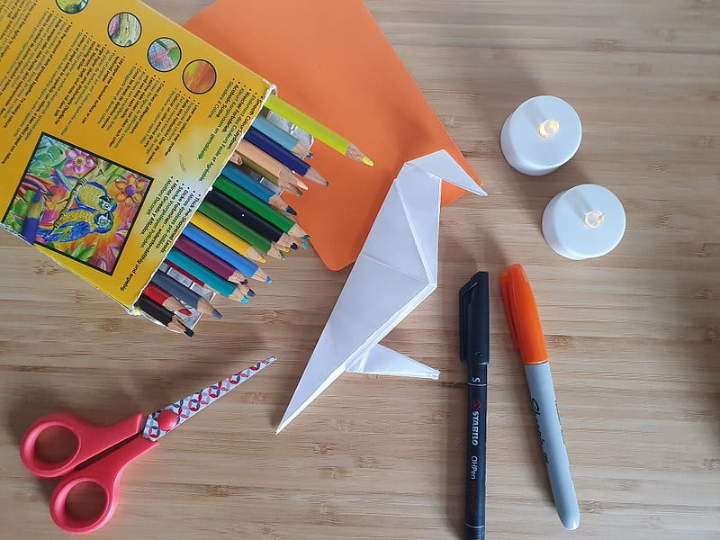White origami crow on the table with pens and colouring pencils next to it.