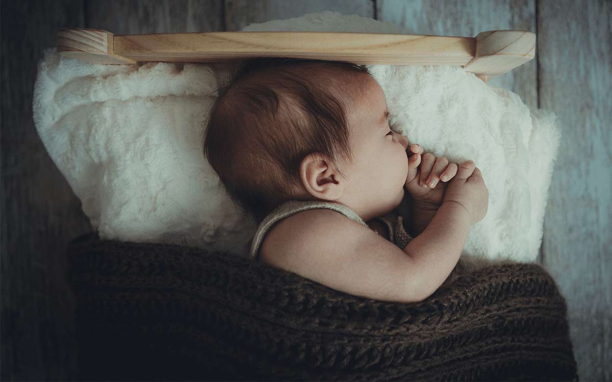 Baby sleeping on its side in a small bed under a brown blanket.