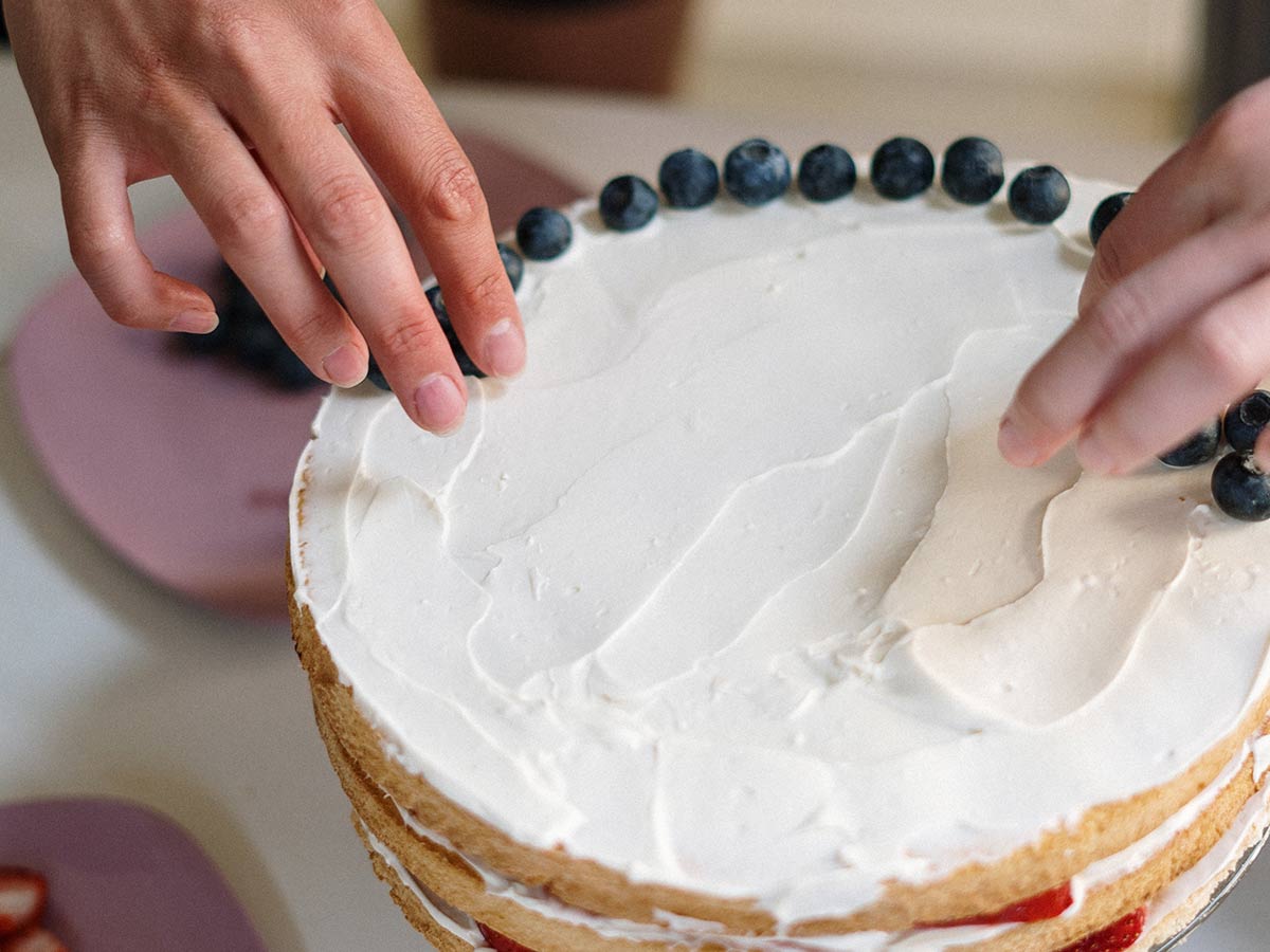 Person decorating an iced cake with blueberries on the top.
