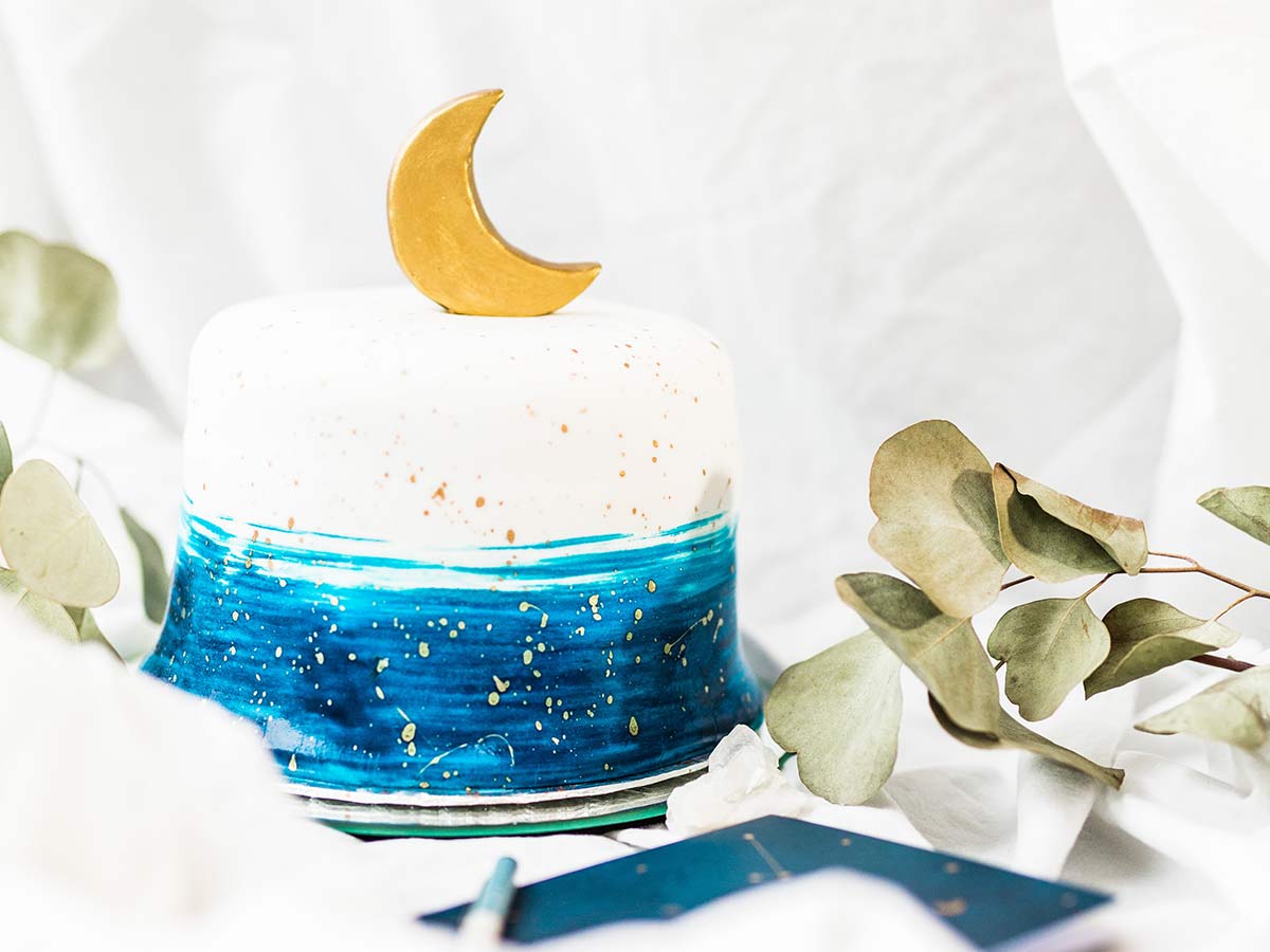White cake with blue icing brushed on around the base, a gold crescent moon on top and gold icing spattering.