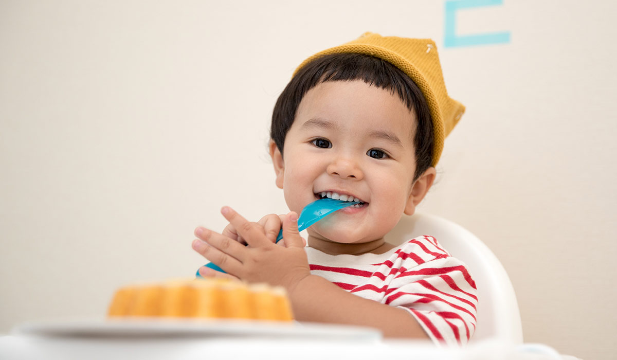 Young boy wearing a knitted yellow crown, sat in his chair eating cake.