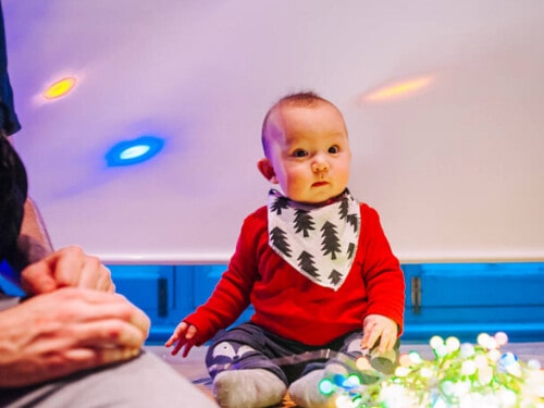 Small baby wearing a bib with Christmas trees on, sat on the floor looking at some Christmas lights.