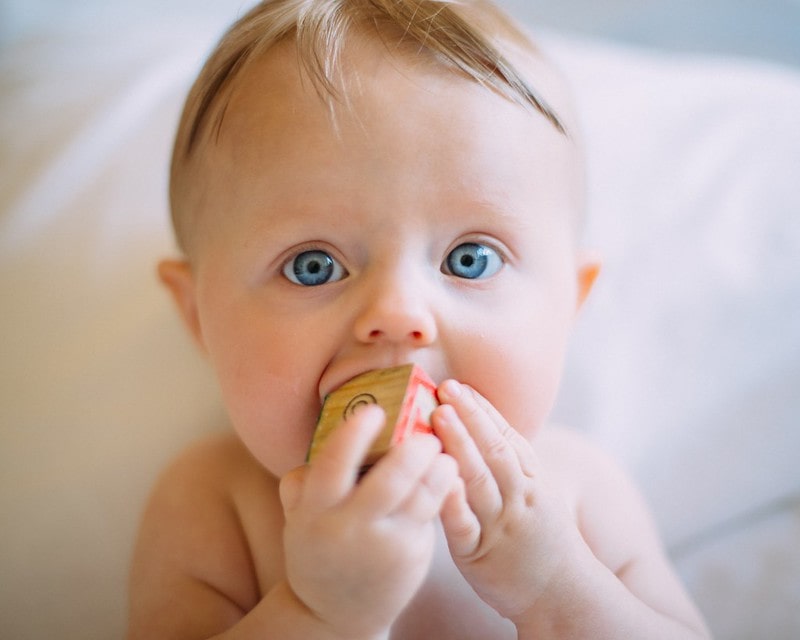 Blue-eyed baby boy with a wooden block in his mouth.