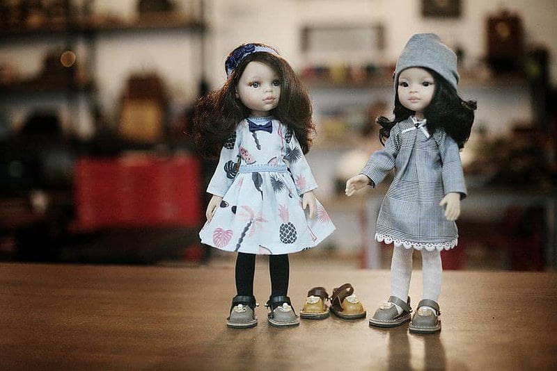Two dolls wearing leather slip-on shoes, with a third pair on the floor between them.