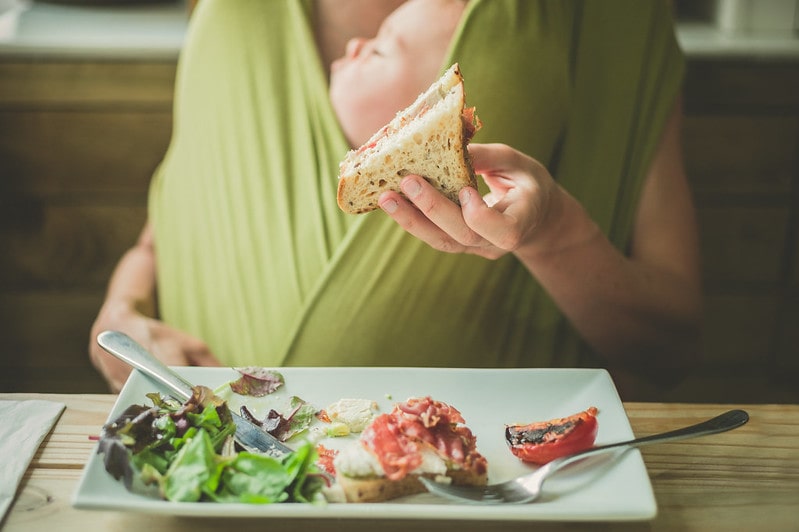 Mum in a restaurant eating a sandwich with her baby in a green baby sling.